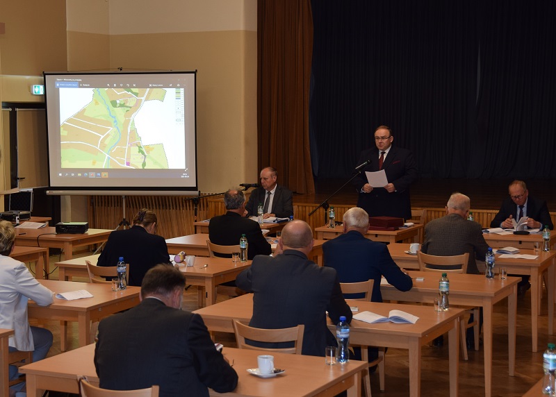 Sesja rady gminy na sali rudzieckiej filii GOK. Wójt przemawia do radnych, którzy siedzą przy osobnych stolikach.